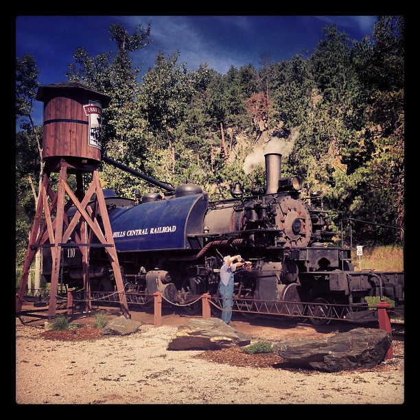 Engineer with oil can oiling a steam locomotive as it sits at a wooden water tower.  
