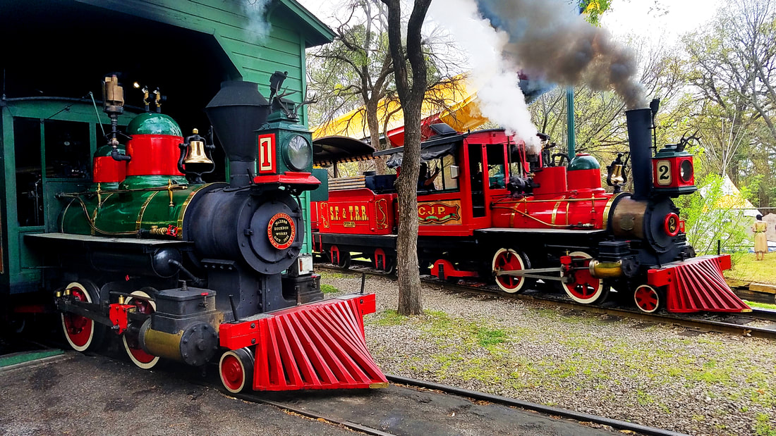 Two Steam locomotives. On the left, a green locomotive with a 1 sits in an engine house. On the right, a red locomotive with a 