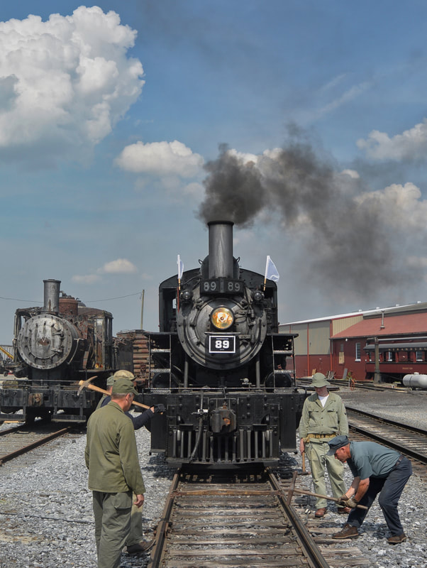 Steam locomotive with #89 and white flags with smoke sits next to an old unused locomotive in a rail yard as a track crew works on the rails with large hammers. 