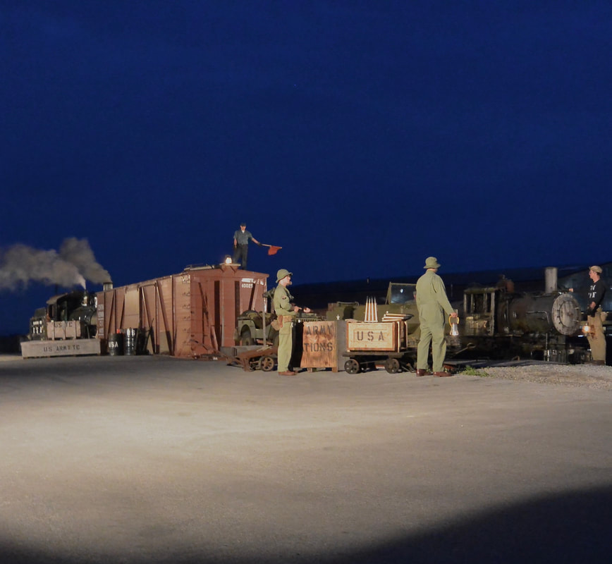 At night, two steam locomotives are visible as well as a boxcar in a rail yard. WWII actors pose with lanterns and a red flag around vintage US Army props. 