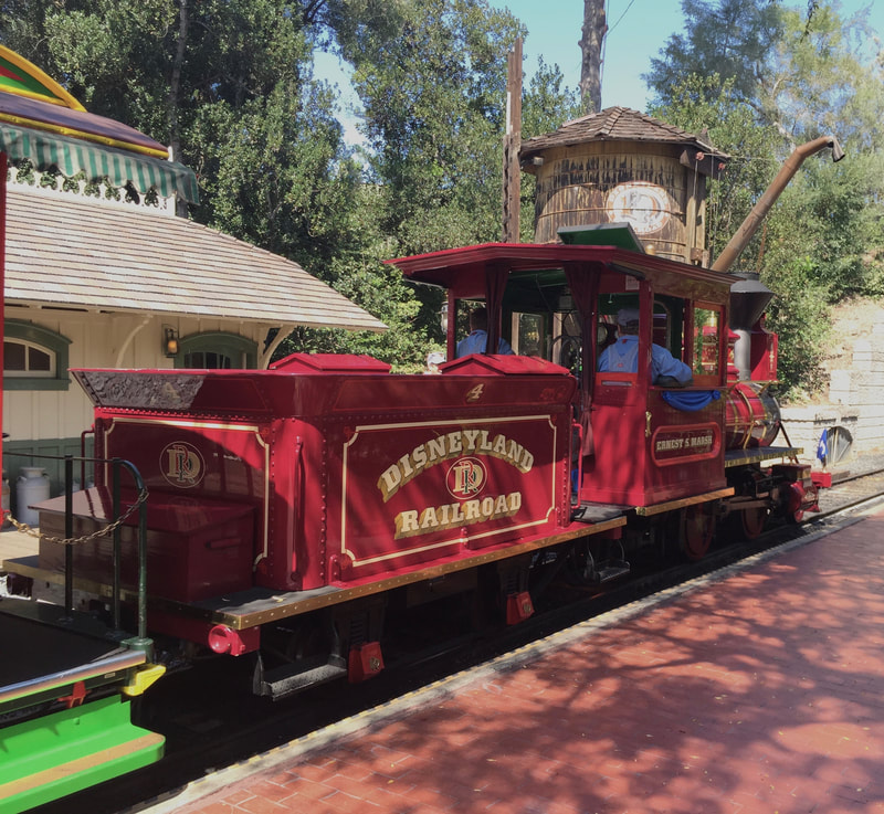 Side of Steam Locomotive arriving on platform with wood water tower and a depot in the background. "Disneyland Railroad 4" is written on the tender. 
