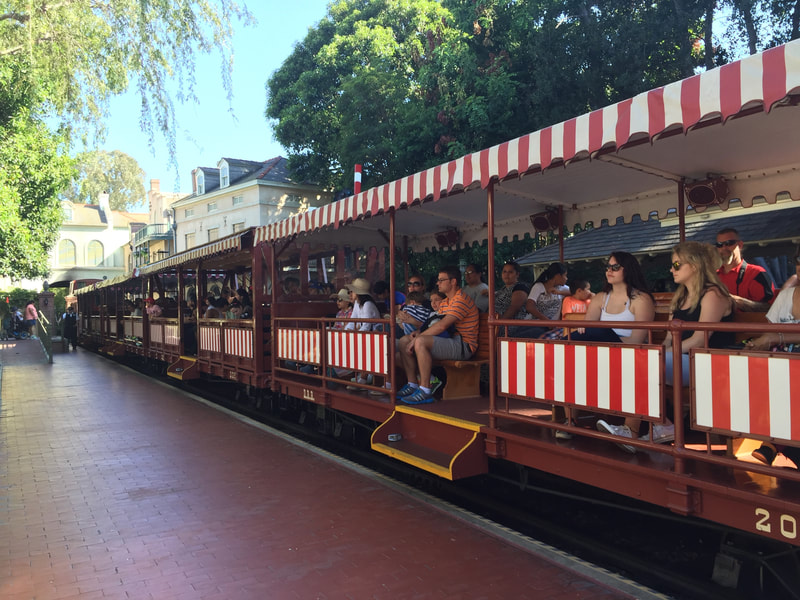Disneyland Passenger train at brick platform. Brown train cars with steps in the middle. Red and White Stripes on roof and sides. 