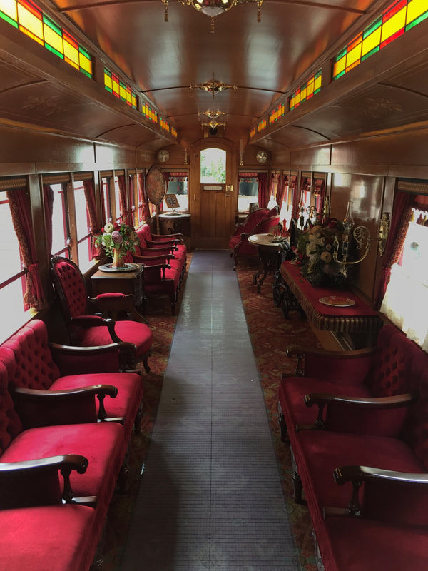 Interior of victorian decorated passenger railcar. Red plush seating, flower vases, and stained glass. 