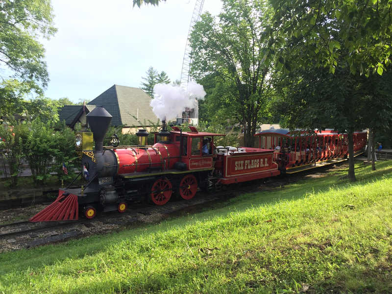 Red Steam Locomotive lets off steam as it pulls train. "Six Flags RR" is written on Tender.