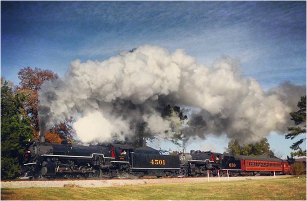 Two Steam Locomotive make steam and smoke as the pull maroon passenger cars. 