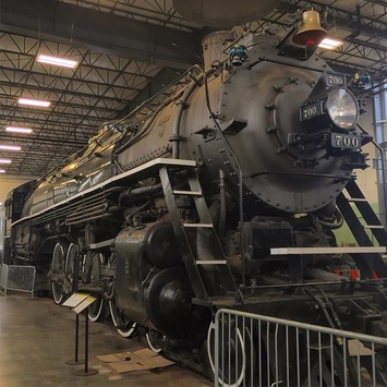 Large Steam locomotive sitting inside a repair shop building. Front of engine has brass bell on top, and headlight in the center. 700 is written in silver below and above the headlight. 