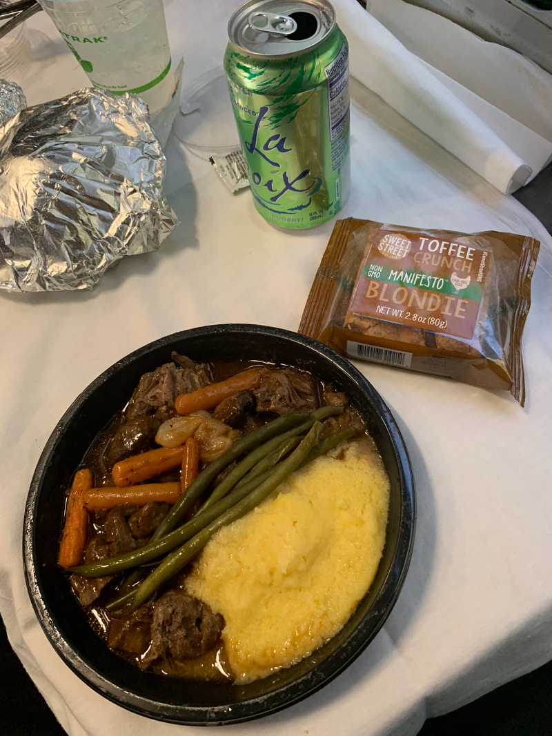 Black plastic bowl with polenta, vegetables and beef sits on a white table with wrapped brownie and can of la croix