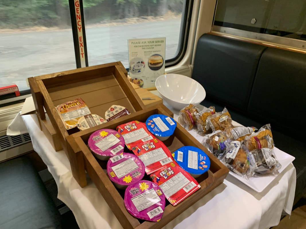 Display of individual servings of cereal, muffins, and fruit bowl on a table inside an Amtrak Dining Car. 