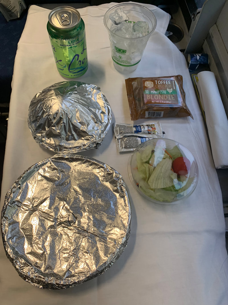 Foil wrapped plates, and small salad in container sit on a white table cloth along with green can of La Croix and plastic wrapped brownie. 
