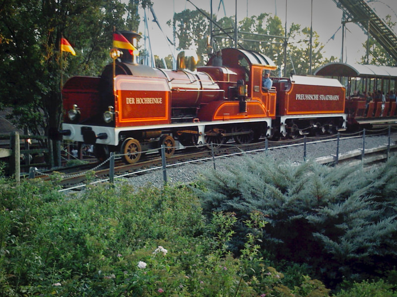 Brown Steam Locomotive with German Flags on the front of the engine pulls a train past a roller coaster. 