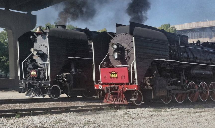Two steam locomotives sit next to each other, both are making smoke. Both have metal smoke deflectors. One has a red front and red wheels. The other has a brass bell. 