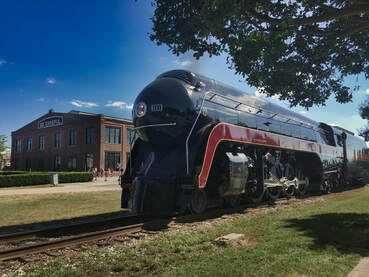 Streamlined Steam locomotive sits on a track in front of an old brick shop building. 