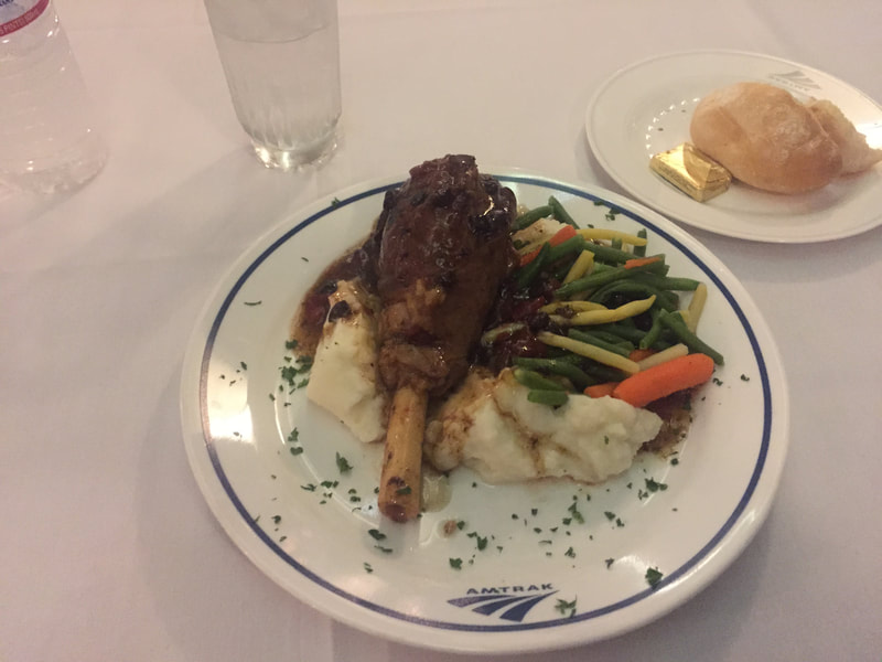Lamb Shank Dinner on an Amtrak Branded China plate sits next to a roll with butter and a glass of ice water on a white table cloth. 