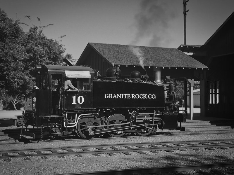 Steam Locomotive making smoke as it moves through rail yard. Engineer is in cab, #10 is written under cab, and "Granite Rock Co." is written on the side of the locomotive. 