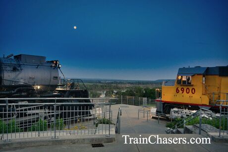 Black Steam Locomotive and Yellow Diesel locomotive on display at park at dusk with full moon overhead. 