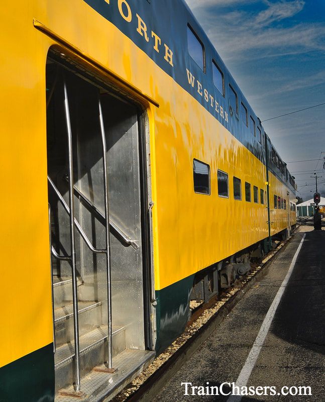 Side view of Green and Yellow Bi-level train car at a station platform. Center door is open with silver steps leading up into the railcar. 