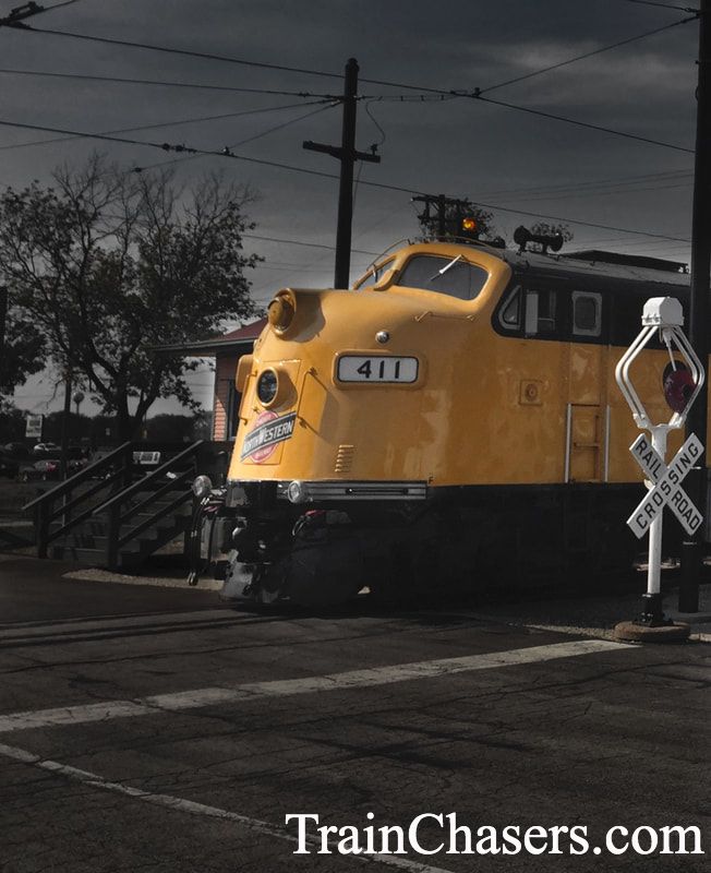 Front of Yellow Diesel locomotive with curved front, 411 is on the locomotive. A old fashioned wig wag railroad crossing signal is at the road crossing. 